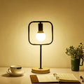 Modern black geometric frame metal table lamp with warm glow, square glass enclosure, and round wooden base for bedside use