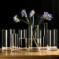 Clear cylindrical glass planter vases of varying heights with subtle vertical ridges, one holding purple flowers on wood coaster