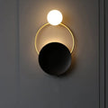 Modern black and gold metal planet wall mount lamp with glowing white spherical bulb and matte black disc bedroom sconce