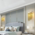 Modern silver and gold tassel design wall lamp with bulb base above a light gray tufted bed with curved headboard and pillows