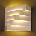Modern white geometric wall mounted lighting sconce with layered stepped design emitting warm ambient light